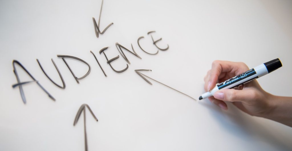 Woman's hand writing the word "audience" on a whiteboard, with arrows.